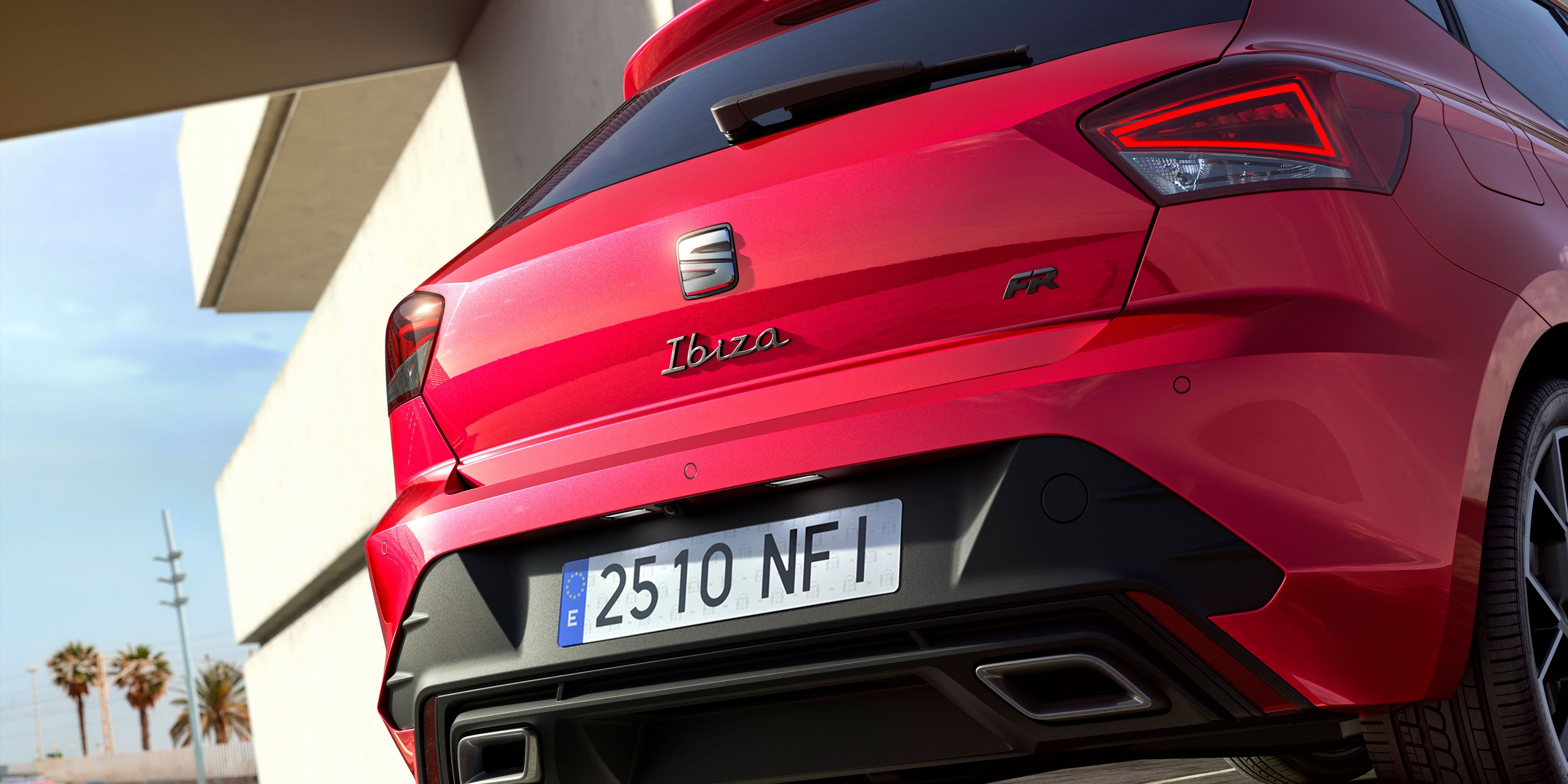 Červené auto značky Seat Ibiza zaparkované v modernom prostredí. Auto je vybavené športovým balíkom FR, čo je viditeľné na jeho výraznom dizajne. V pozadí sú palmy a jasná obloha.