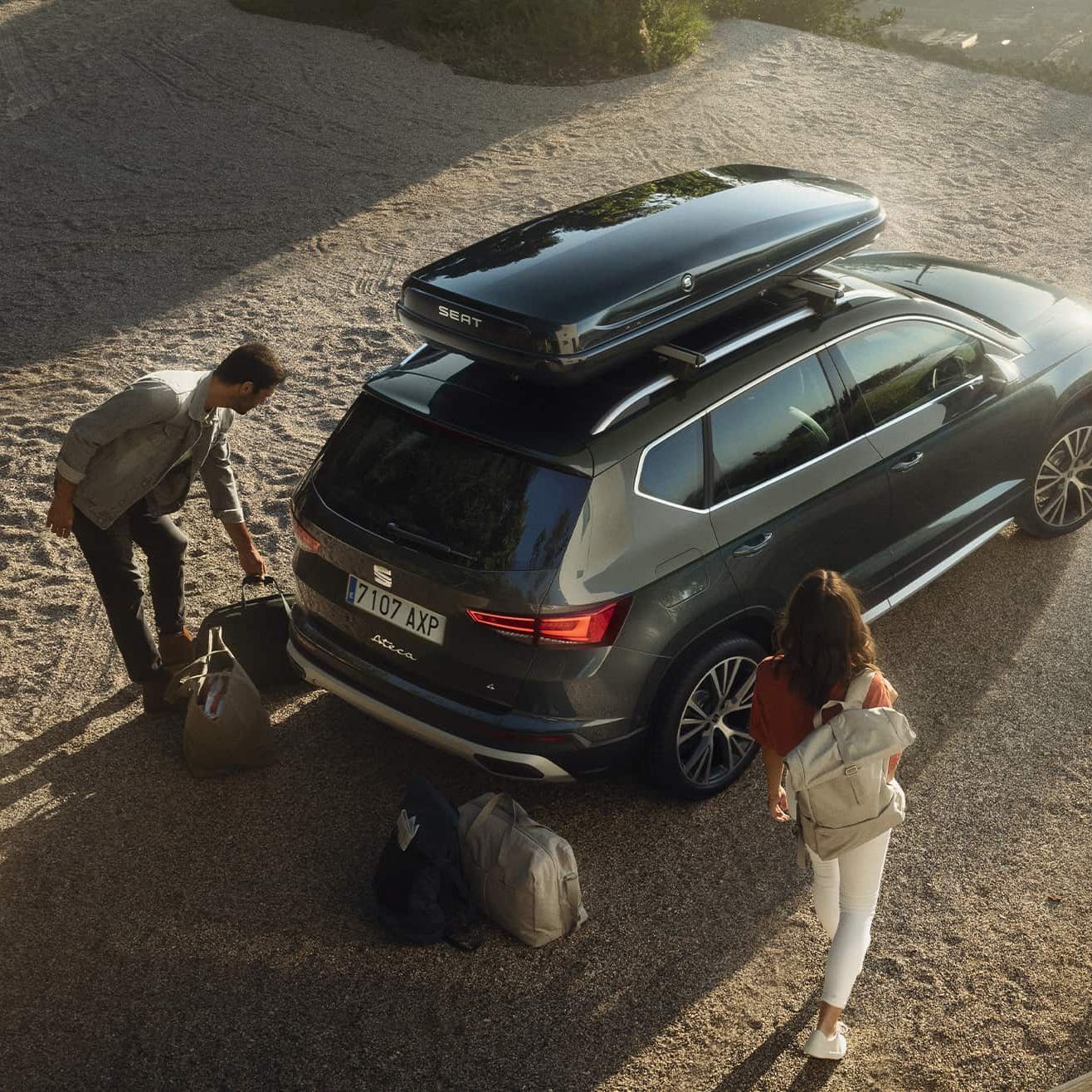 Pohľad zhora na SEAT Ateca so strešným boxom. Pri vozidle sú položené cestovné tašky. Muž a žena prichádzajú k vozidlu.