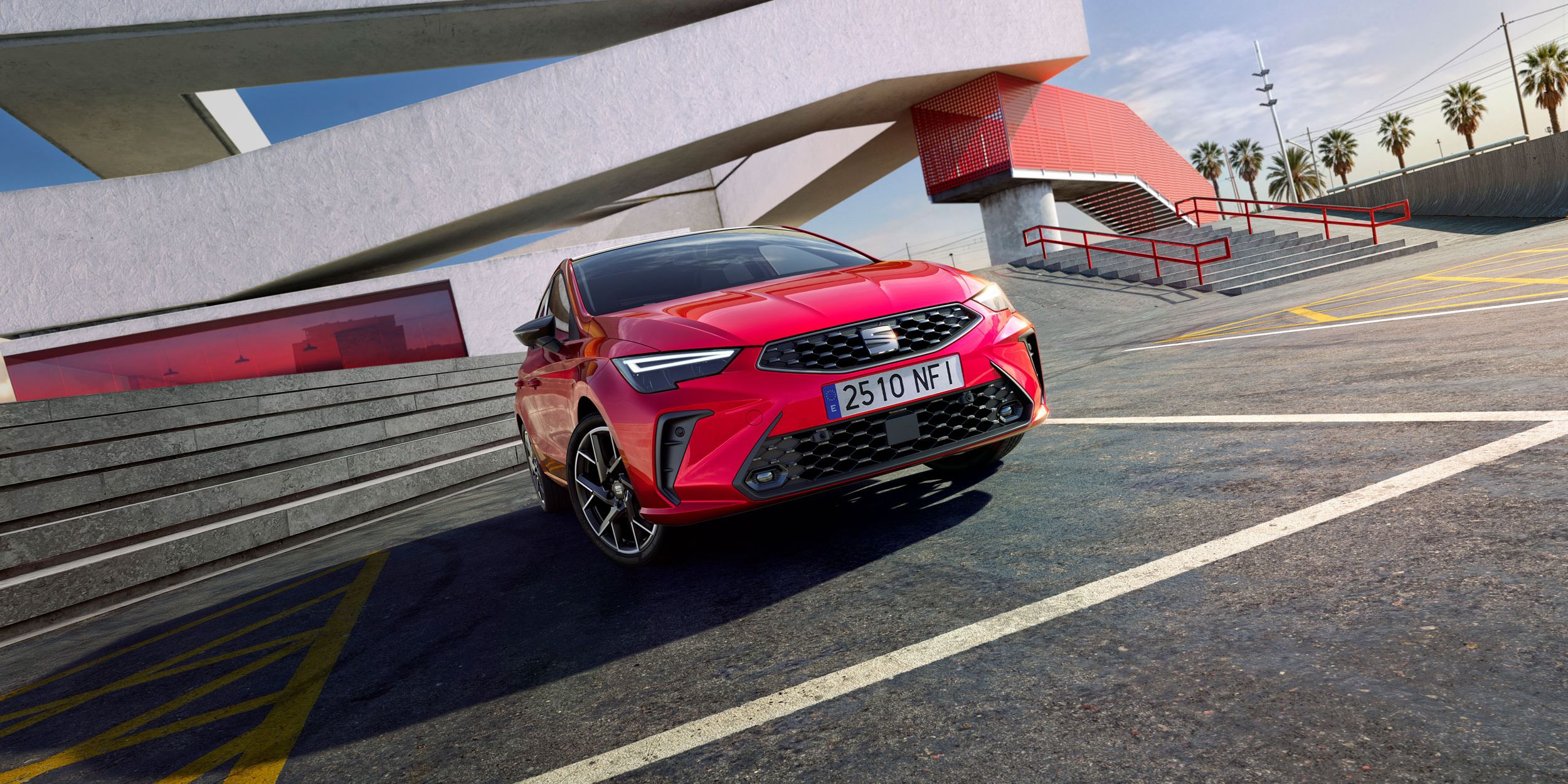 Červený SEAT Ibiza vedľa moderného architektonického objektu s veľkými geometrickými líniami. Na pozadí sú palmy a modrá obloha. Auto má španielsku poznávaciu značku.