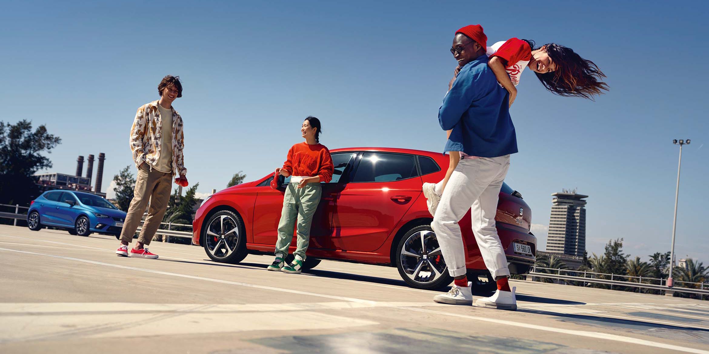 Dva vozidlá na parkovisku. O červené auto sa opiera mladá žena. Vedľa stojí mladý muž a ďalší dvaja sa zabávajú o niečo ďalej.