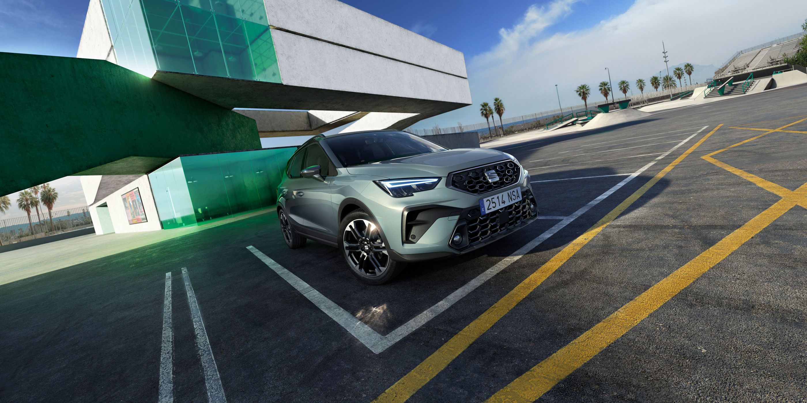 Automobil stojí na parkovisku pred modernou budovou zo skla a betónu. Obloha je slnečná, na horizonte vidieť oblačnosť. V pozadí sú palmy.