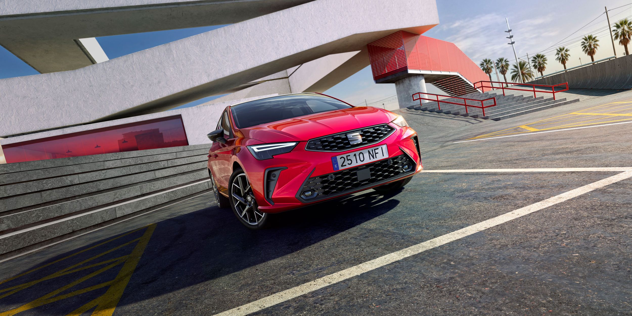 Červený SEAT Ibiza vedľa moderného architektonického objektu s veľkými geometrickými líniami. Na pozadí sú palmy a modrá obloha. Auto má španielsku poznávaciu značku.