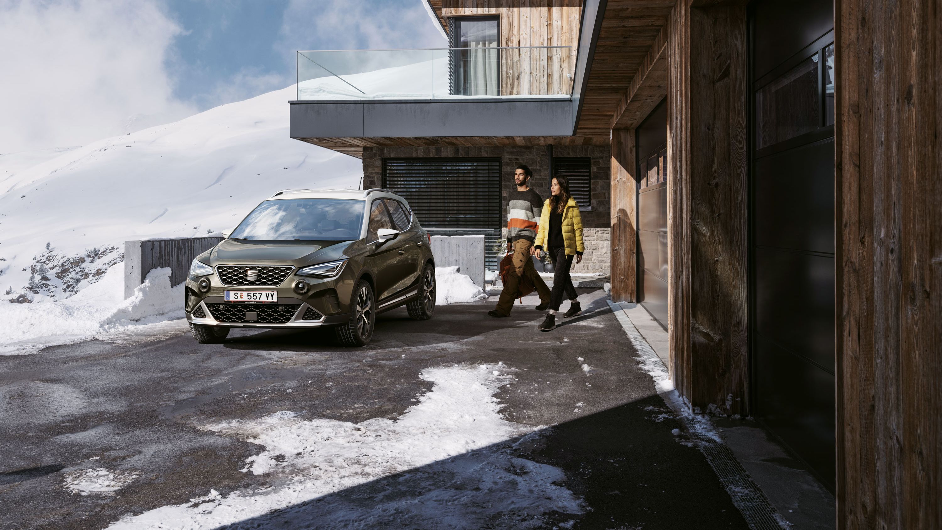 SEAT Arona stojí pri drevenom, modernom dome. Muž a žena kráčajú k vozidlu.