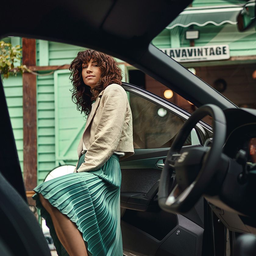 Mladá žena v zelenej skladanej sukni stojí vo tvorených dverách vozidla a nohu má vyloženú na prahu dverí. Pozerá sa priamo do objektívu. Interiér vozidla je rozostrený.