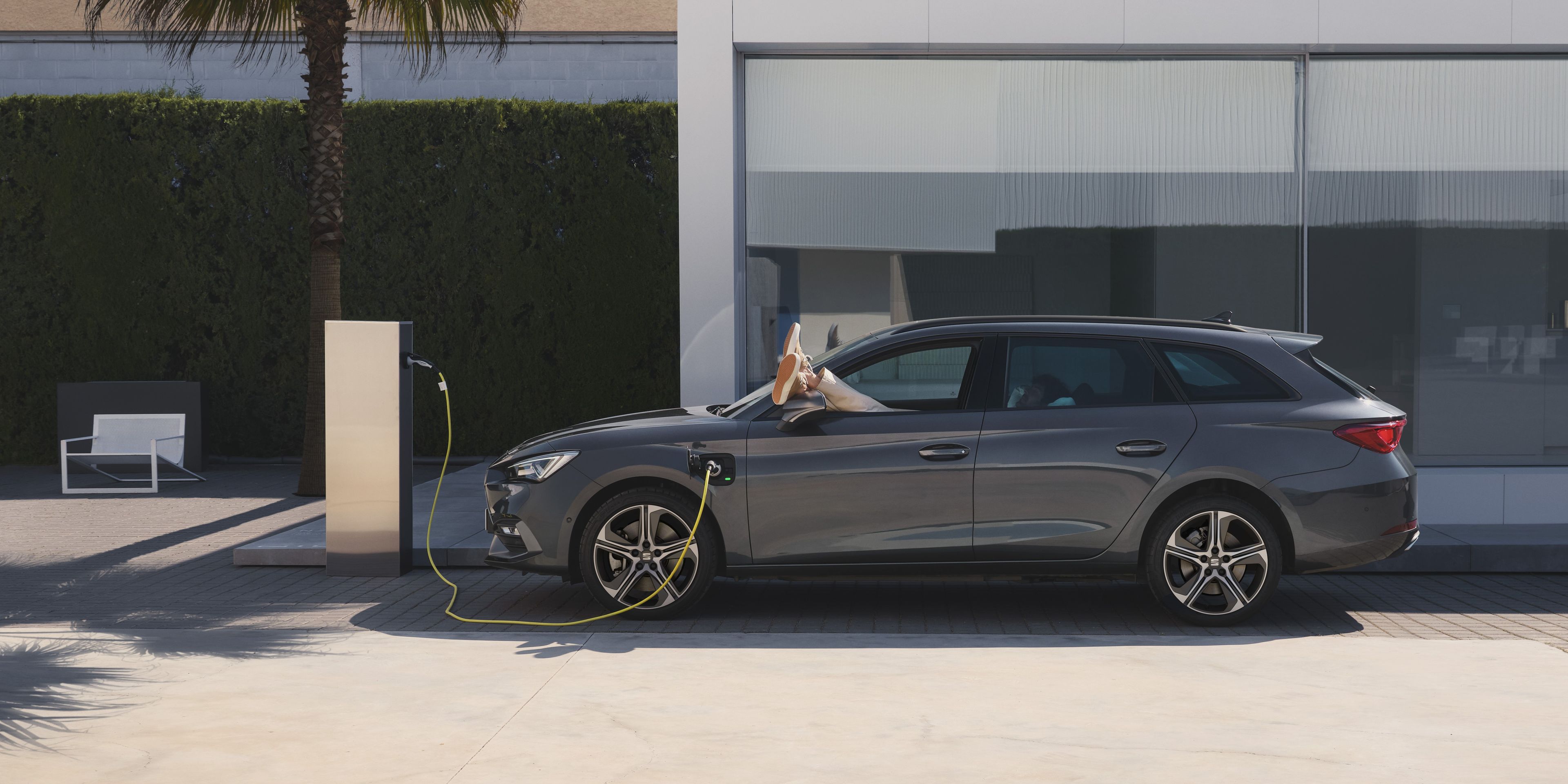 SEAT Leon Sportstourer, v ktorom leží muž na sedadle vodiča a má vyložené nohy von z okna, sa nabíja na wallboxe pred moderným domom s veľkými sklenenými oknami.