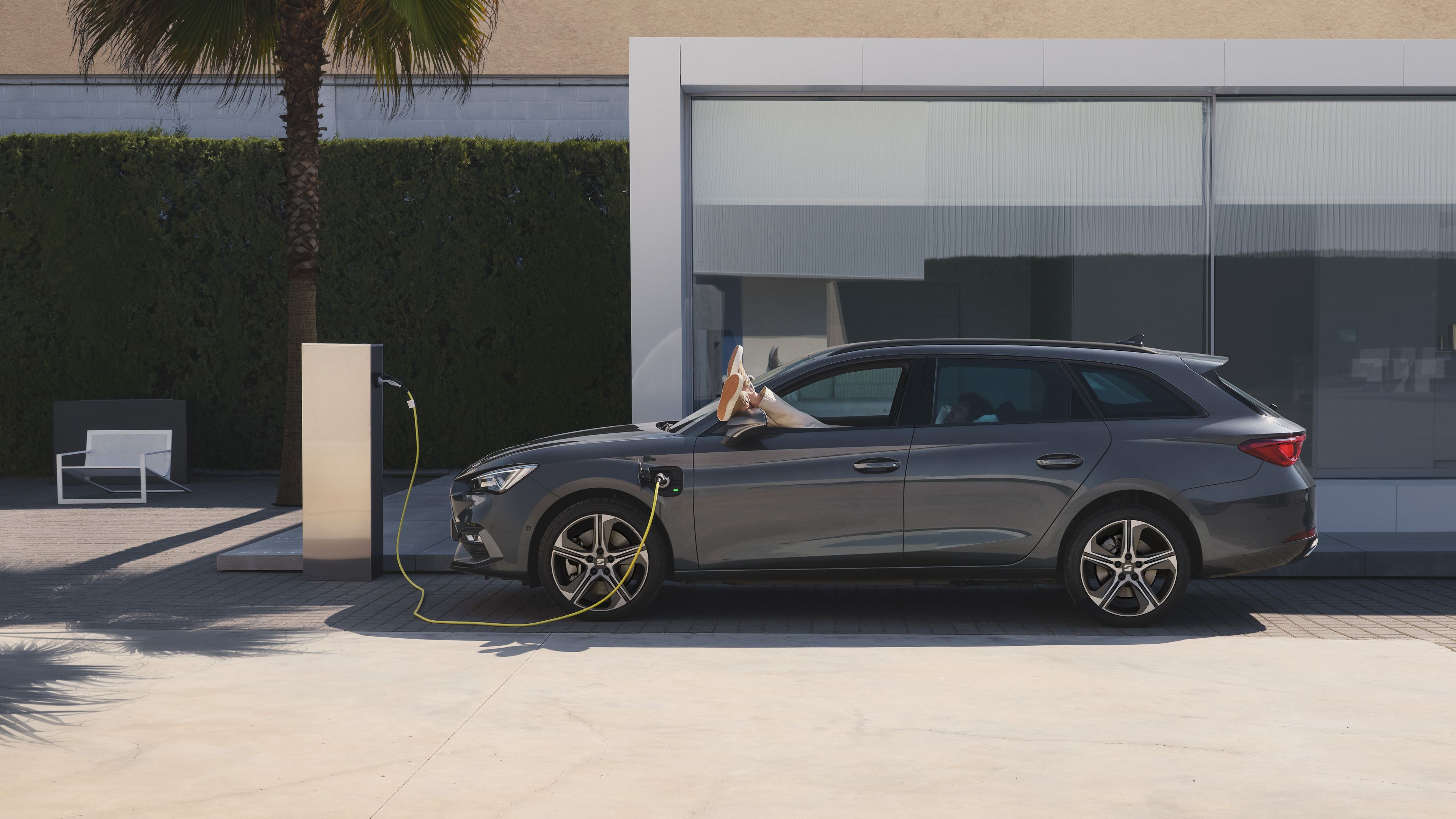 SEAT Leon Sportstourer, v ktorom leží muž na sedadle vodiča a má vyložené nohy von z okna, sa nabíja na wallboxe pred moderným domom s veľkými sklenenými oknami.