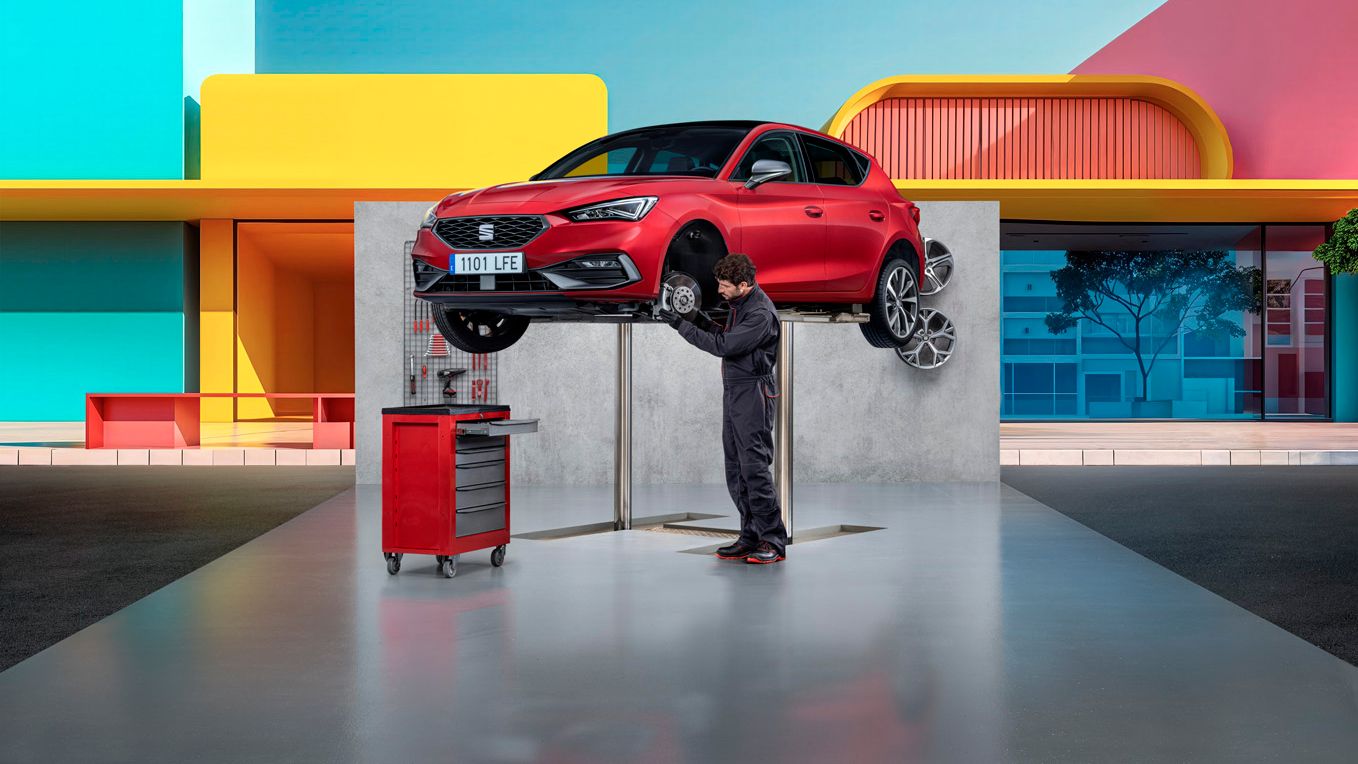 SEAT Ibiza na zdviháku vo výške. Servisný pracovník stojí pri vozidle a kontroluje brzdový kotúč. V pozadí sú pestrofarebné domy.