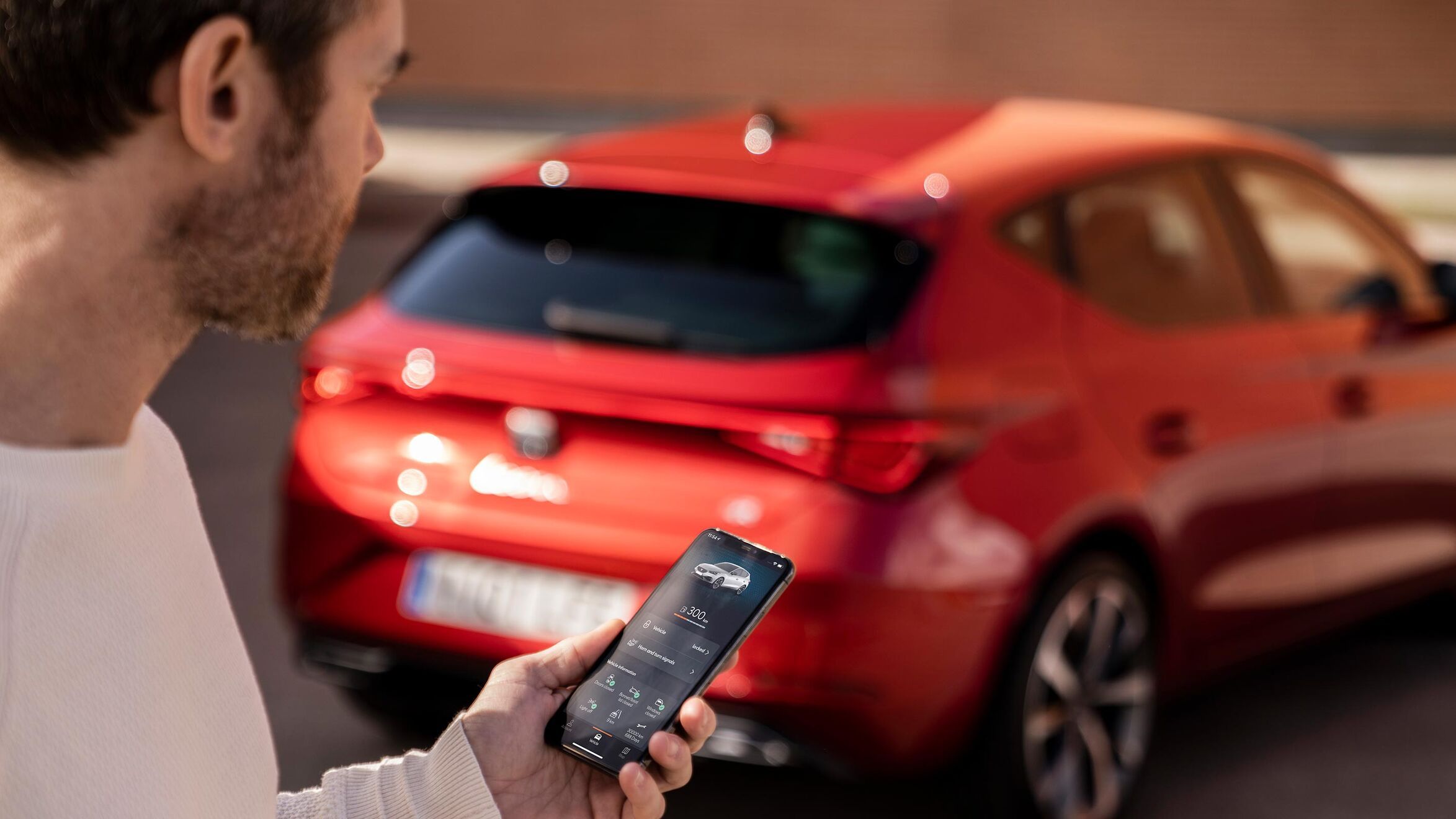 Muž so smartfónom v ruke, na ktorého displeji sú zobrazené rôzne údaje o vozidle. V pozadí je rozostrený SEAT Leon.