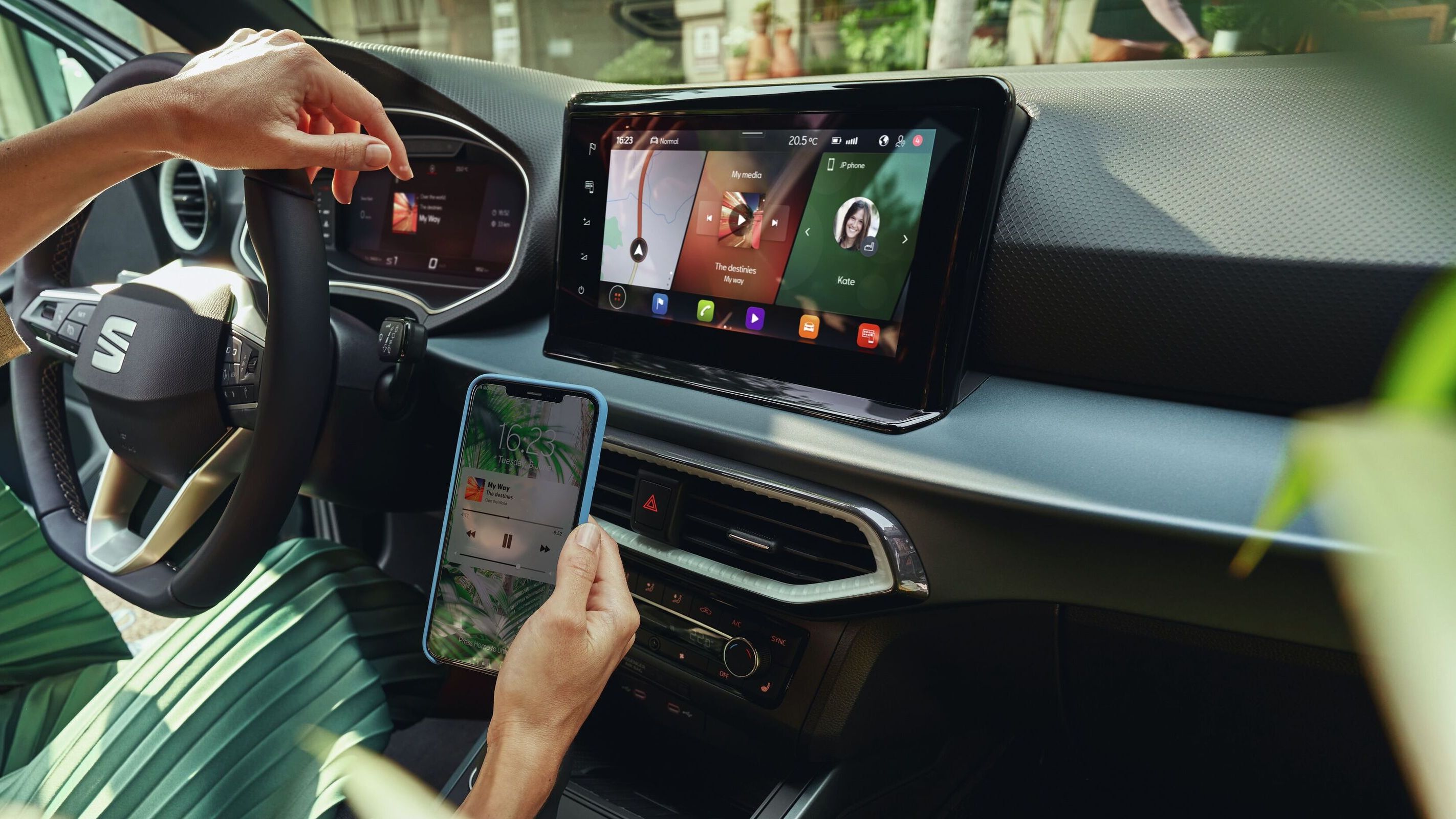 Žena na sedadle vodiča sa jednou rukou opiera o multifunkčný volant a v druhej ruke drží smartfón s hudobnou aplikáciou. Vidieť palubnú dosku so všetkým, čo zahŕňa. 