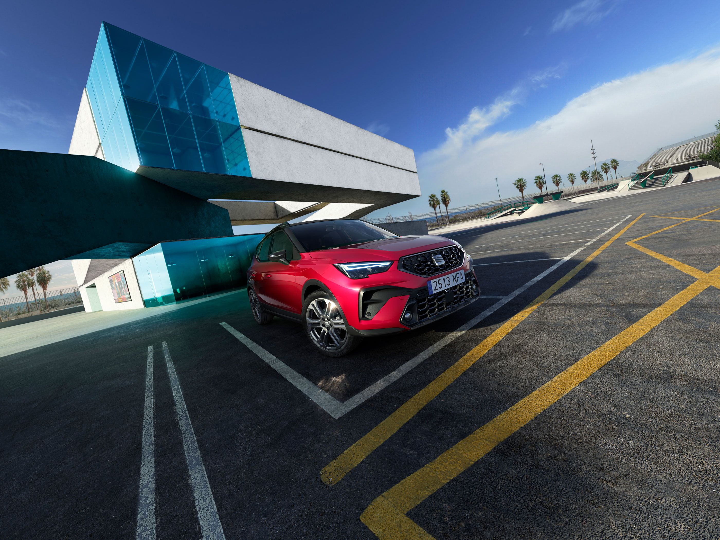 Červené SUV zaparkované pred modernou budovou so sklenenými a betónovými prvkami. Na pozadí sú viditeľné palmy a modrá obloha.