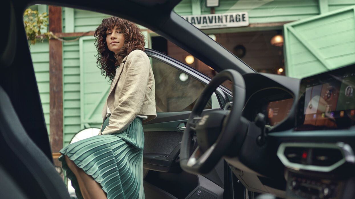 Mladá žena stojí v otvorených dverách vodiča automobilu, ktorý stojí pred kaviarňou. Fotené je to z miesta spolujazdca a vidieť aj časť interiéru.