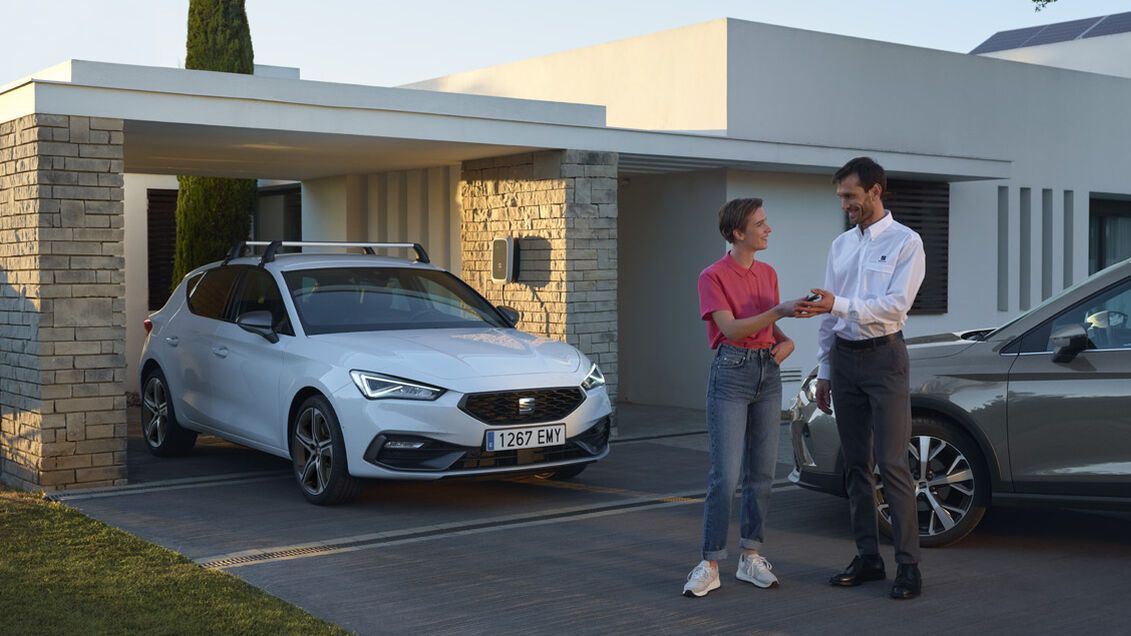 SEAT Leon zaparkovaný pod prístreškom pri modernej vile. Pred vozidlom stoja dvaja ľudia a za nimi je ďalšie auto.