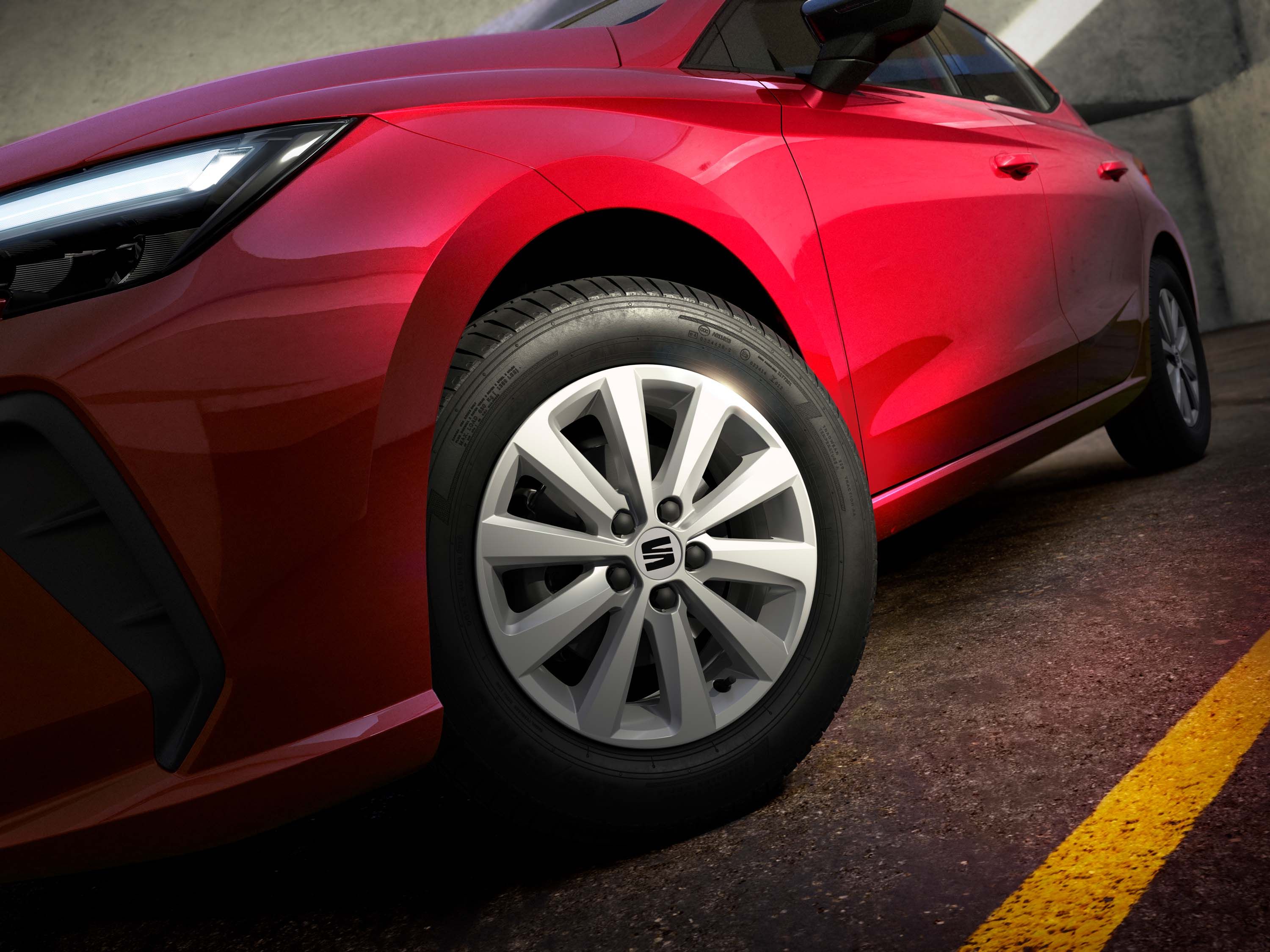 Detailný záber na červený automobil, zameraný na jeho predné koleso. V pozadí je viditeľný betónový múr. Automobil stojí na asfaltovej ceste.