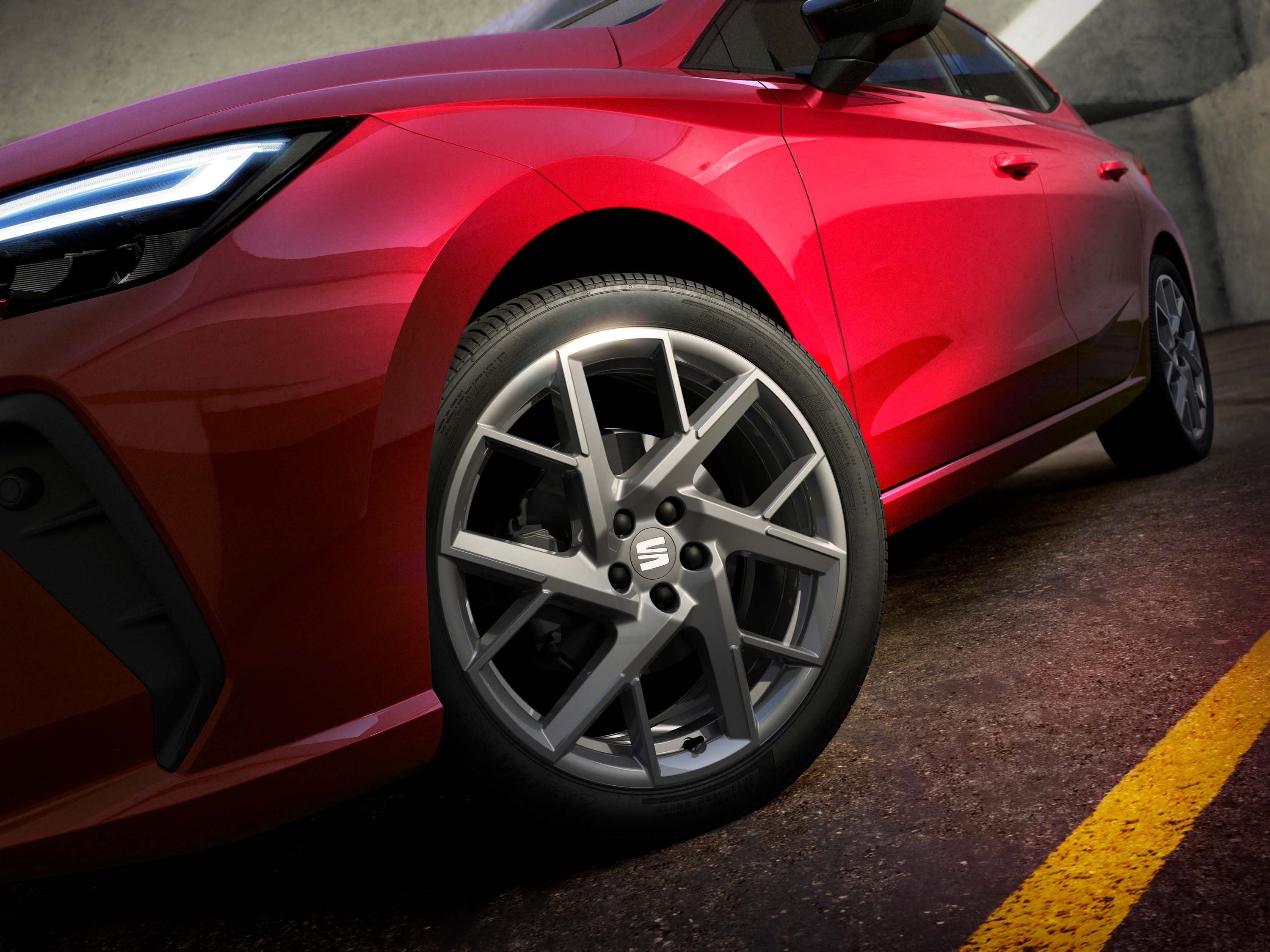 Červené auto s detailom na predné koleso. Koleso má hliníkové disky s viacnásobnými lúčmi. Auto je zaparkované na asfaltovom povrchu.