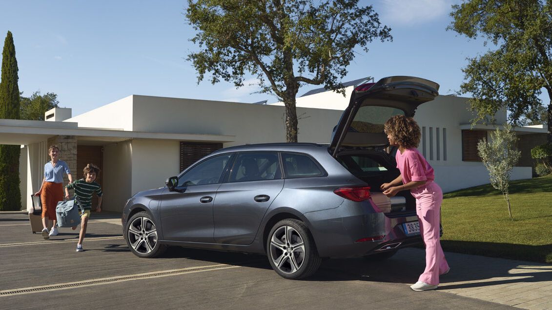 Žena stojí pri vozidle SEAT Leon s otvorenými dverami batožinového priestoru a čaká na chlapca a ženu, ktorí idú smerom k nej. Vozidlo je zaparkované pri ceste pred modernou budovou.