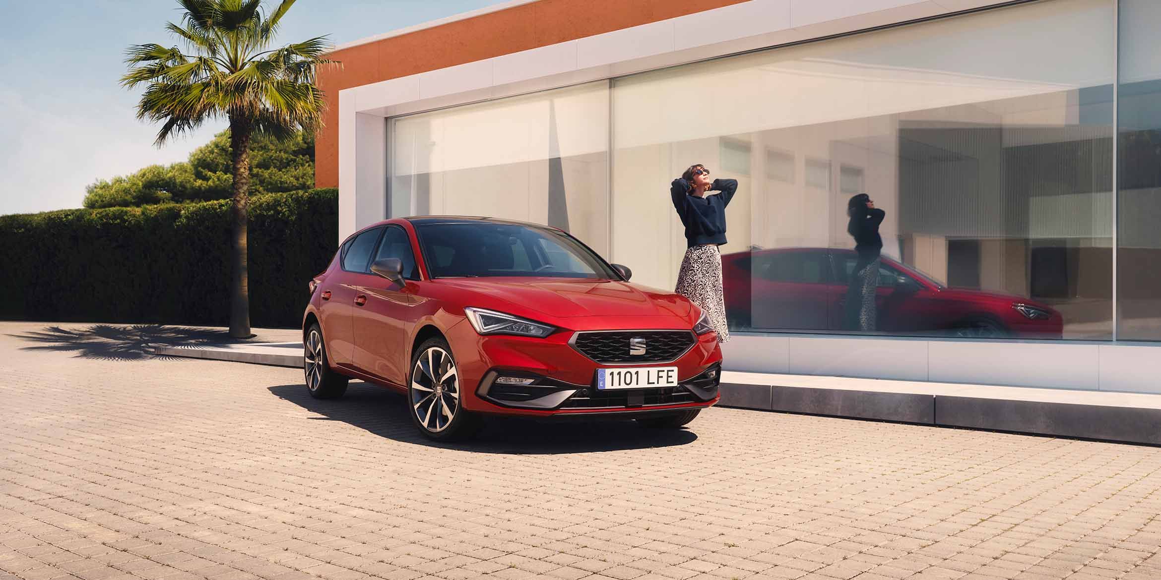 Červený SEAT Leon, pri ktorom stojí žena je zaparkovaný pre moderným domom s veľkými sklenenými oknami, palmou a plotom z kríkov.