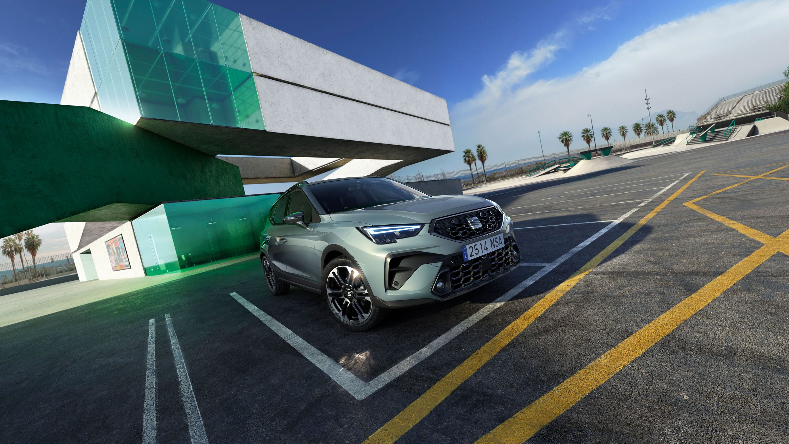 Automobil stojí na parkovisku pred modernou budovou zo skla a betónu. Obloha je slnečná, na horizonte vidieť oblačnosť. V pozadí sú palmy.