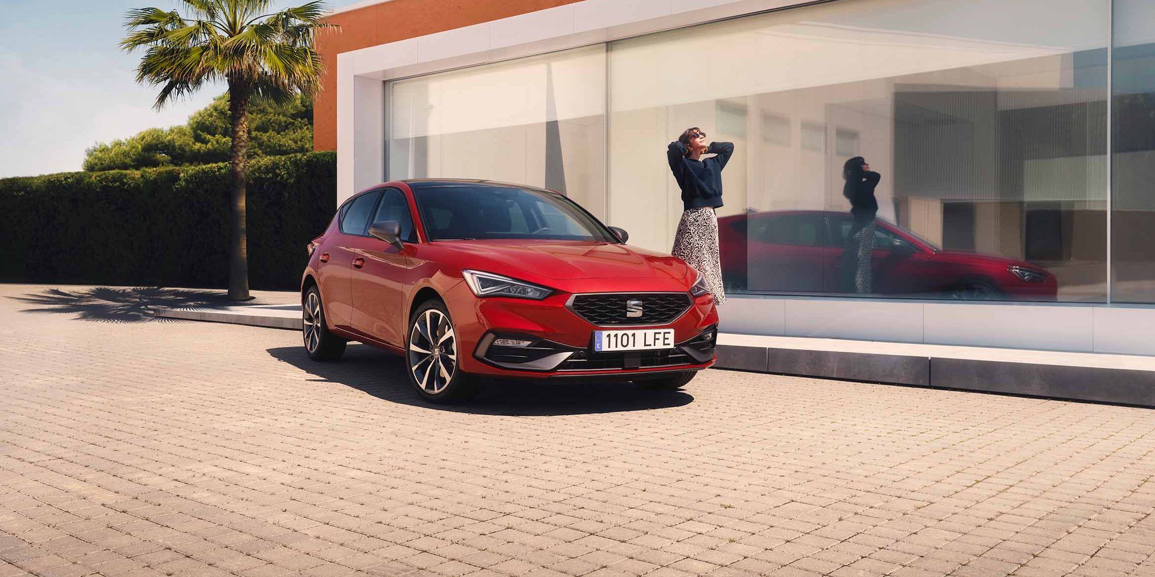 Červený SEAT Leon, pri ktorom stojí žena je zaparkovaný pre moderným domom s veľkými sklenenými oknami, palmou a plotom z kríkov.