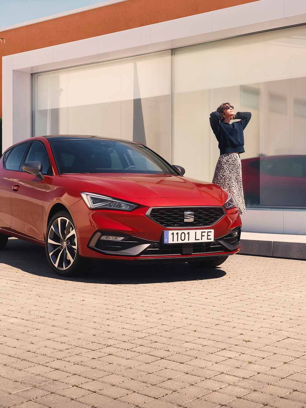 Červený SEAT Leon, pri ktorom stojí žena je zaparkovaný pre moderným domom s veľkými sklenenými oknami, palmou a plotom z kríkov.