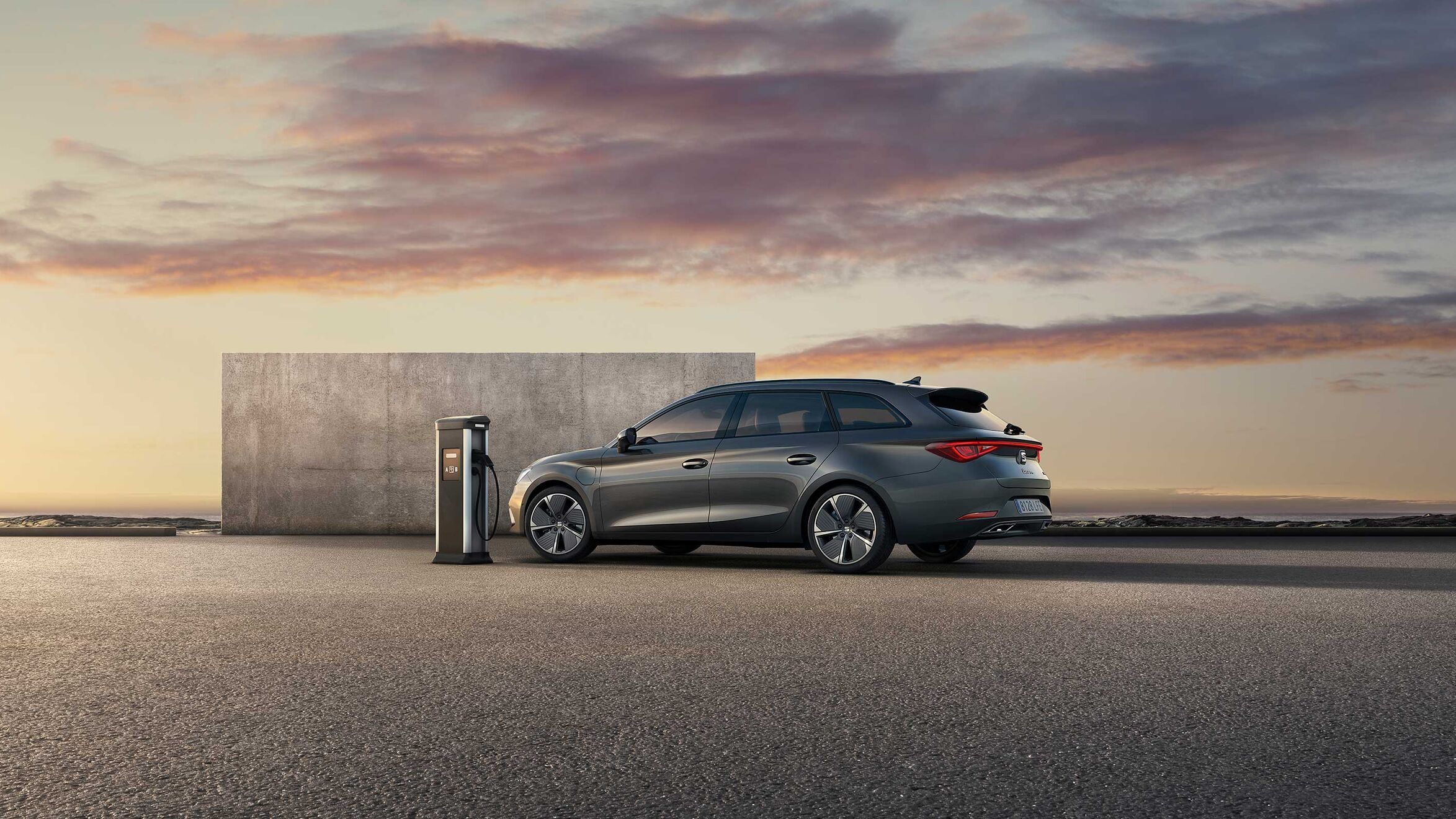 SEAT Leon Sportstourer eHybrid na voľnej ploche pri betónovom múre a verejnej nabíjacej stanici. V pozadí čiastočne vidieť hory. Scénu dotvára zafarbenie neba pri západe slnka.