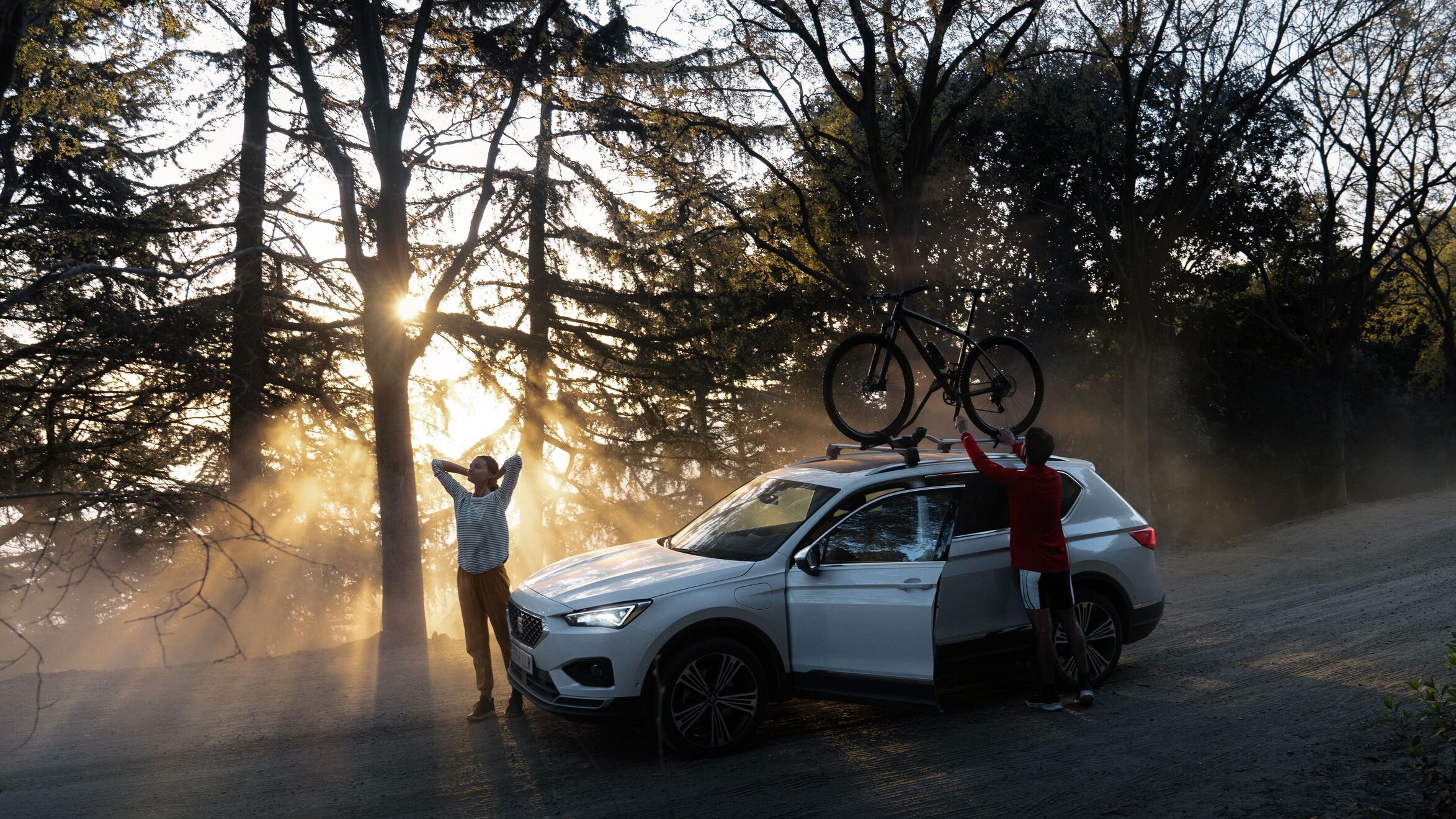 SEAT Ateca stojí na lesnej ceste. Žena stojí pred vozidlom a naťahuje sa, muž skladá zo strešného nosiča horský bicykel. Scénu dotvárajú slnečné lúče prenikajúce spoza stromu.