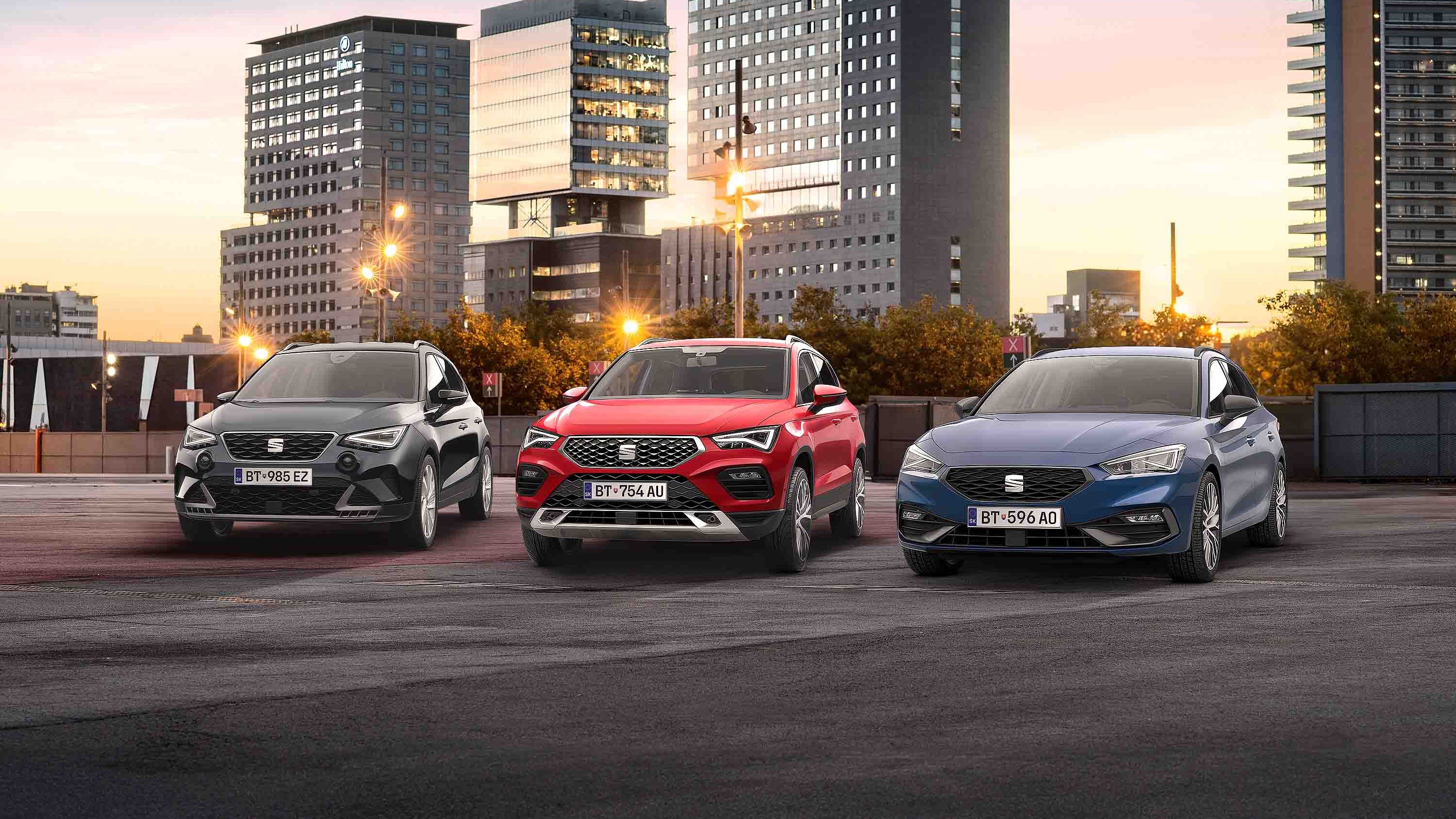 SEAT Leon, Arona a Ateca stoja na parkovisku Za nimi vidieť vysoké mestské budovy a časť mesta pri západe slnka.