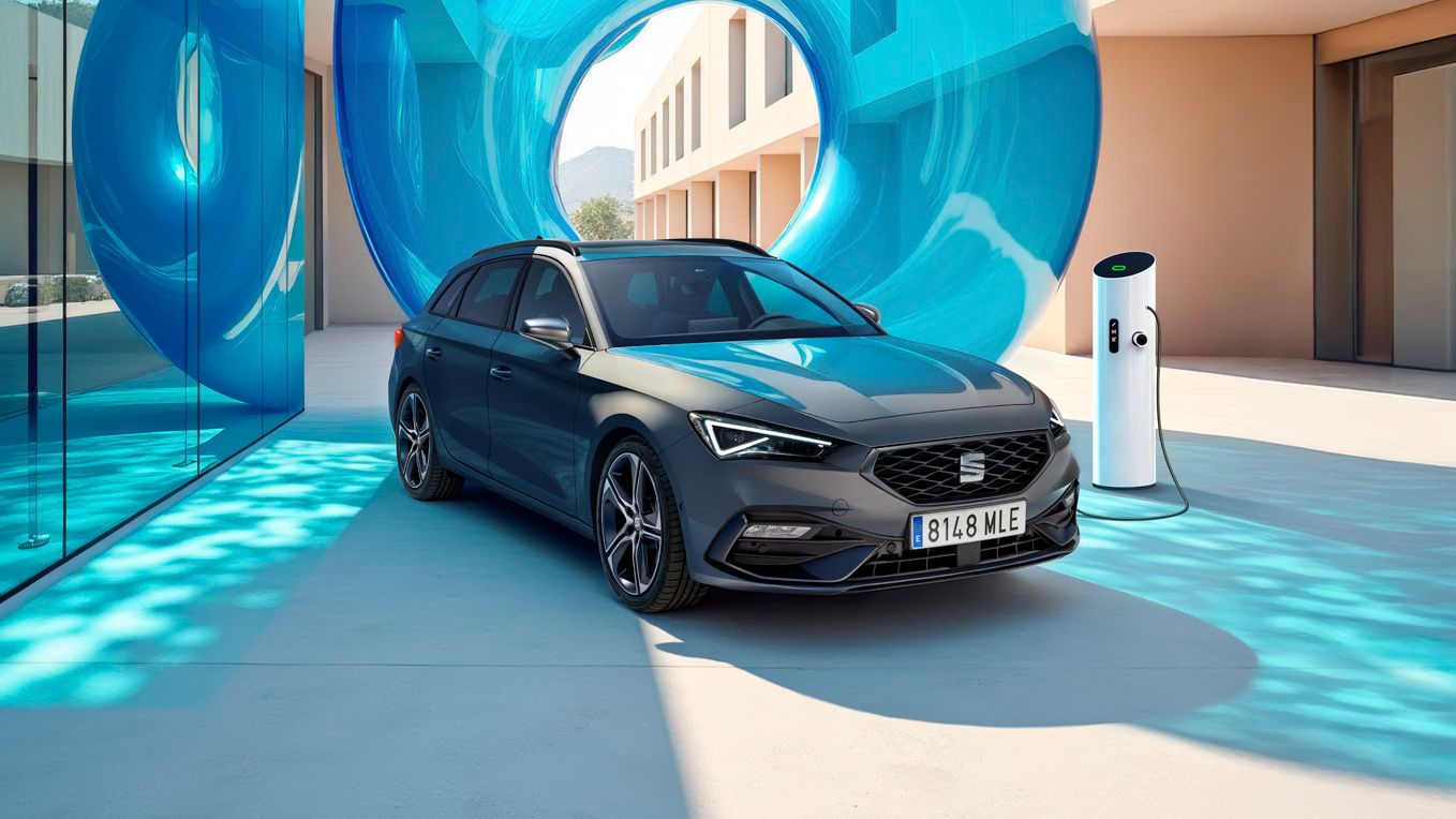 SEAT Leon sa nabíja pri modernej bytovke z jednej strany s presklenou stenou a modrou priehľadnou inštaláciou za ním.