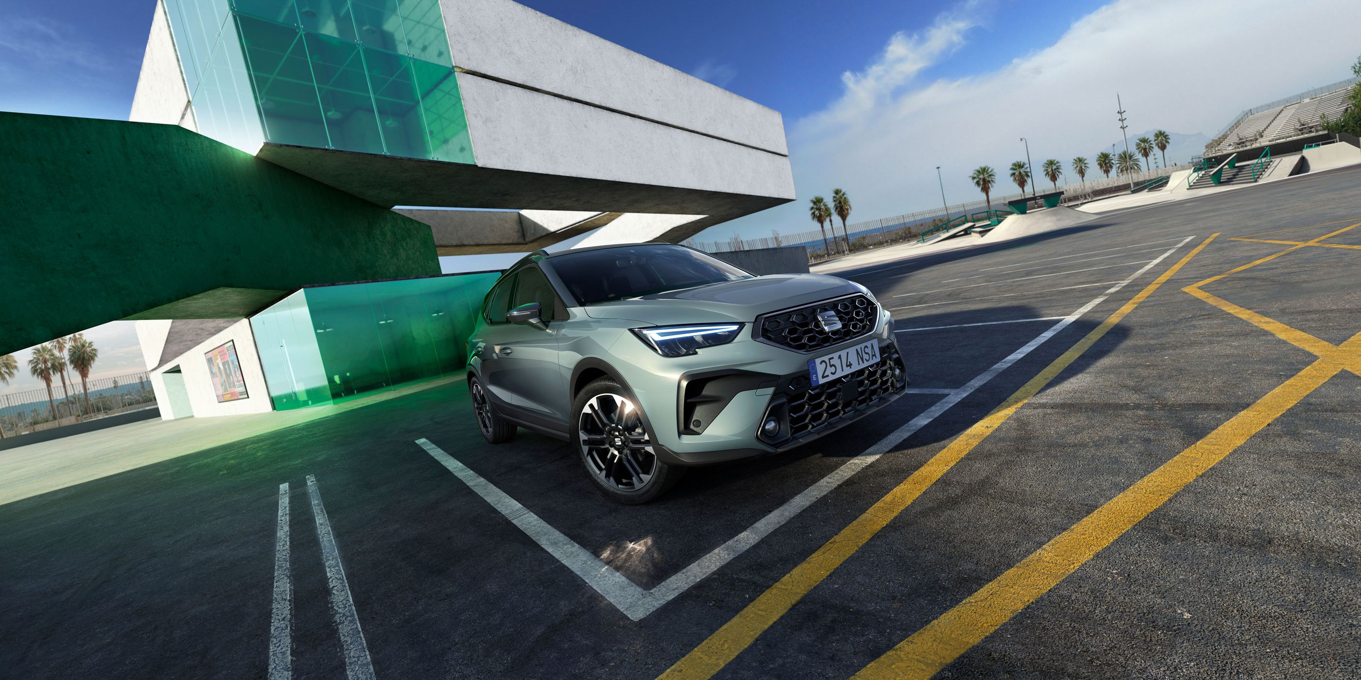 Automobil stojí na parkovisku pred modernou budovou zo skla a betónu. Obloha je slnečná, na horizonte vidieť oblačnosť. V pozadí sú palmy.