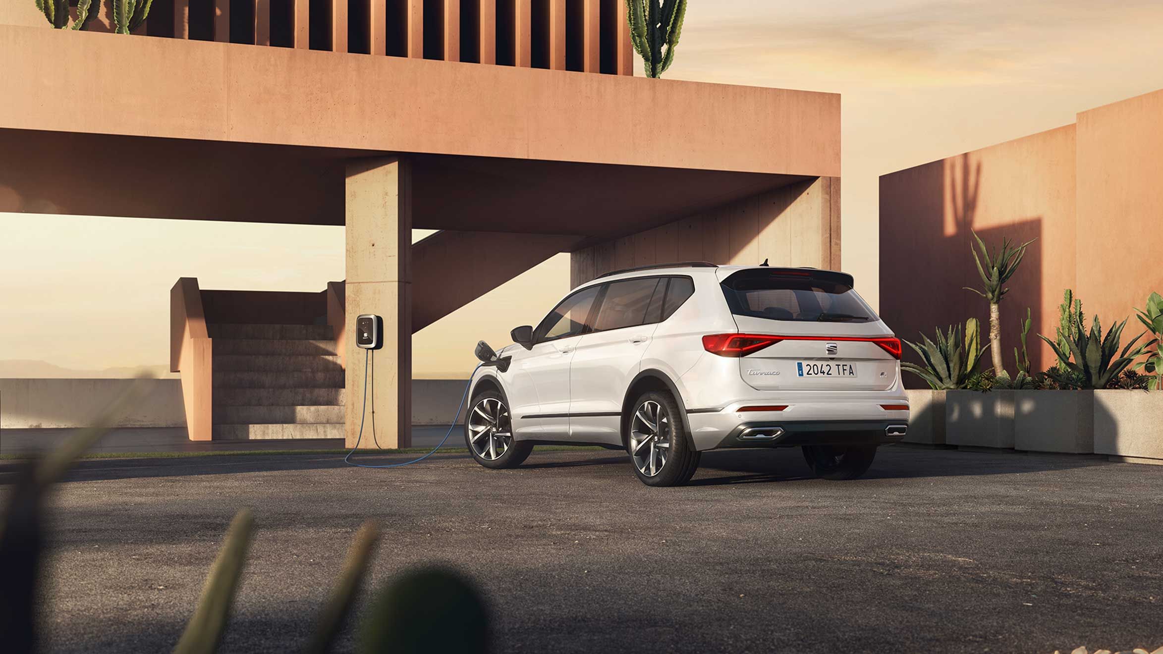 SEAT Tarraco FR PHEV pri modernej budove s domácou nabíjacou stanicou, ku ktorej je napojený a nabíja sa. Okolo je veľa zelene.