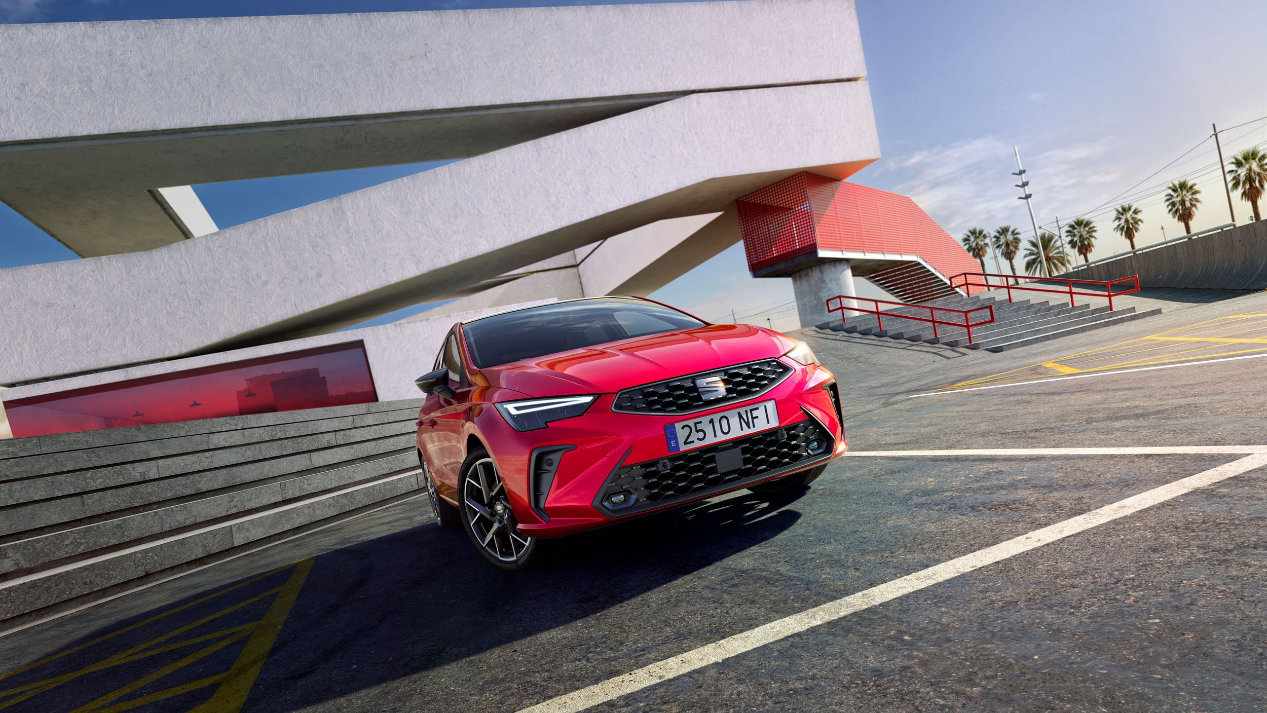 Červený SEAT Ibiza vedľa moderného architektonického objektu s veľkými geometrickými líniami. Na pozadí sú palmy a modrá obloha. Auto má španielsku poznávaciu značku.