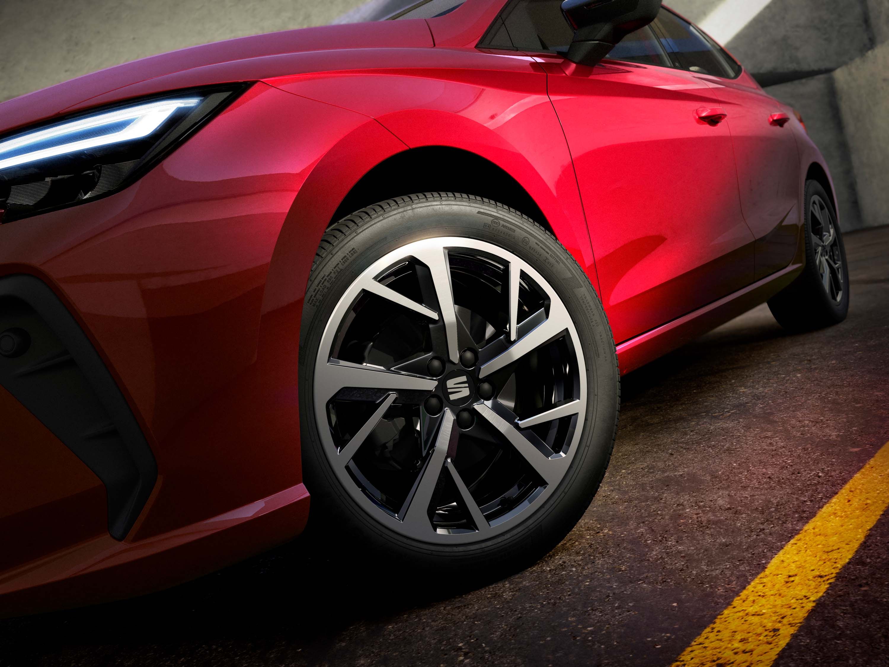 Červené športové vozidlo zaparkované na ceste s moderným dizajnom kolies a svetlometov. Predná polovica a kolesá sú detailne zobrazené. Auto stojí na asfaltovom povrchu s viditeľnou čiarou.