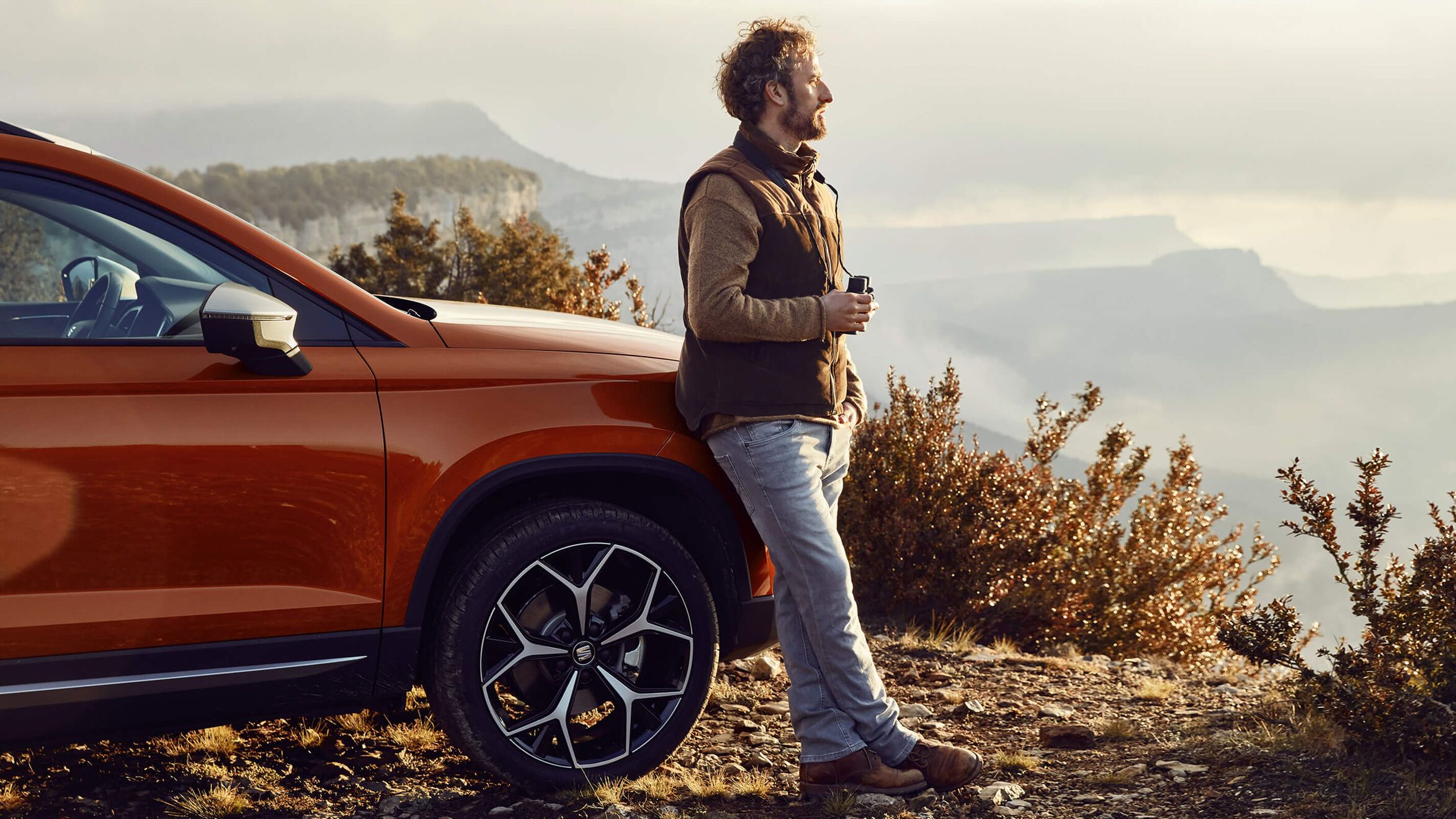 Muž sa opiera o vozidlo SEAT. V ruke drží kávu a užíva si výhľad na okolie.