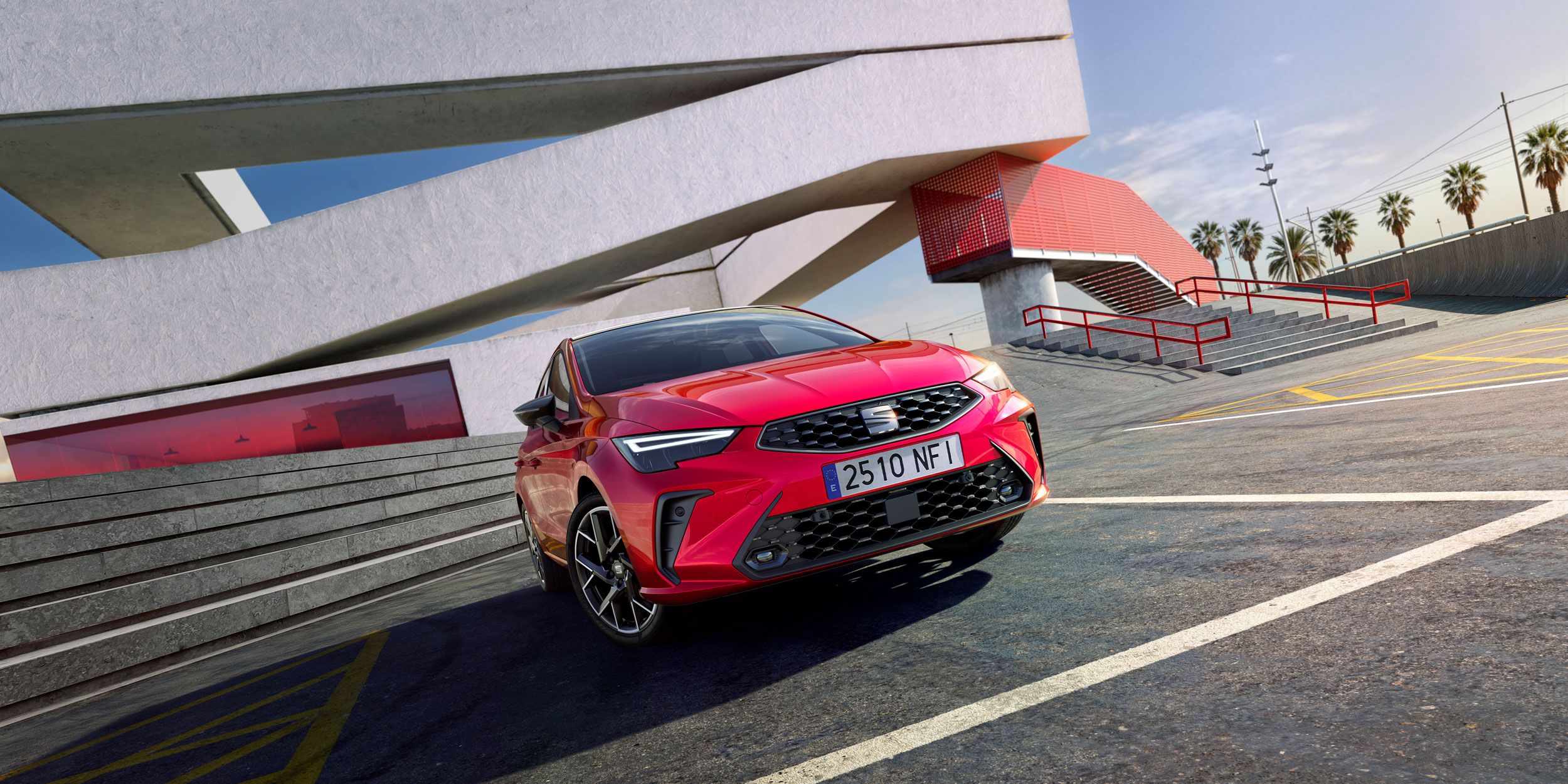 Červený SEAT Ibiza vedľa moderného architektonického objektu s veľkými geometrickými líniami. Na pozadí sú palmy a modrá obloha. Auto má španielsku poznávaciu značku.