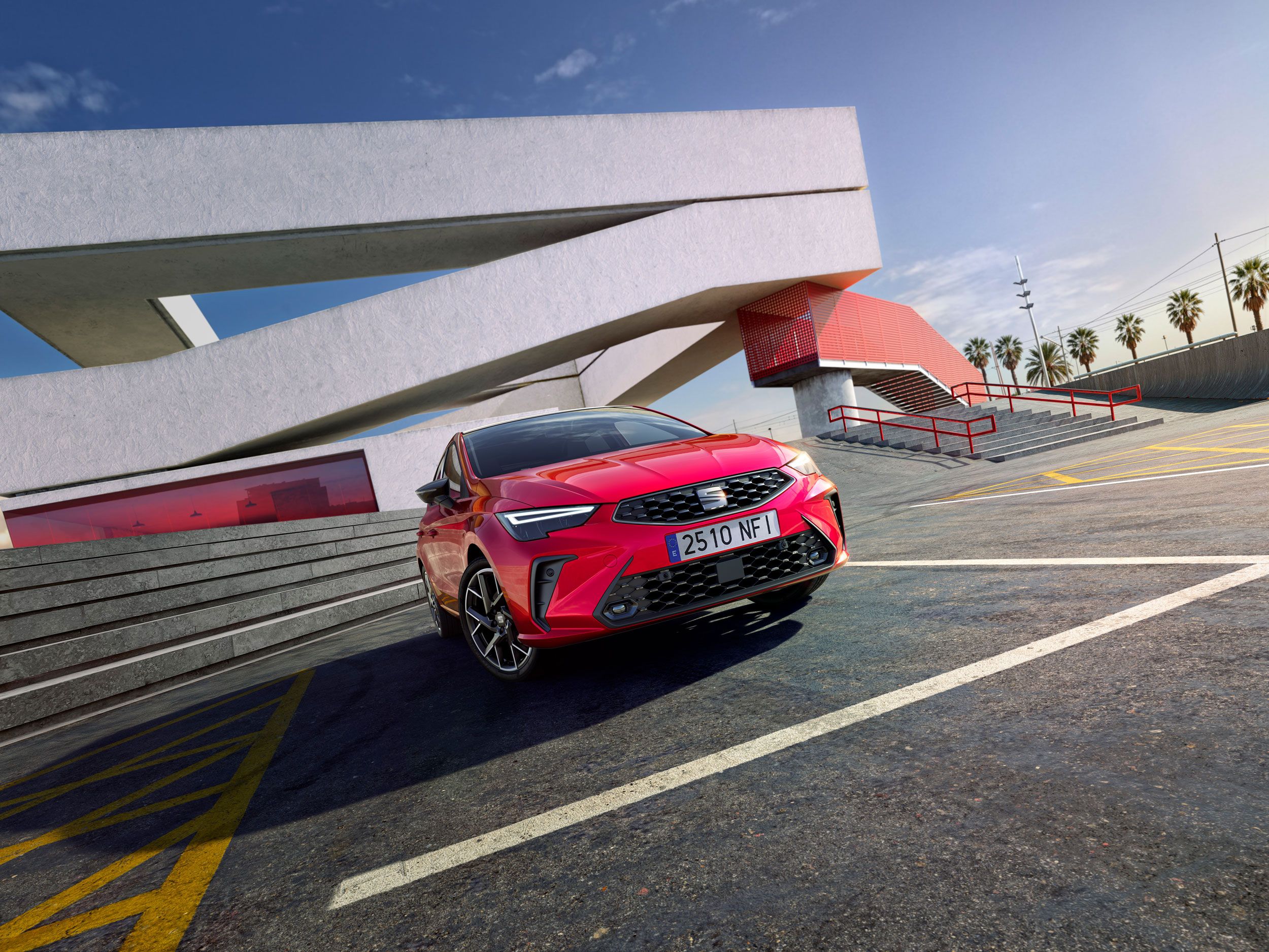 Červený SEAT Ibiza vedľa moderného architektonického objektu s veľkými geometrickými líniami. Na pozadí sú palmy a modrá obloha. Auto má španielsku poznávaciu značku.